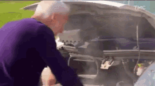 a man in a purple sweater is looking under the hood of a car with smoke coming out of it .