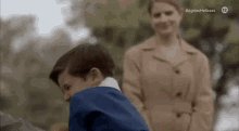 a little boy in a blue suit is standing next to a woman in a brown coat .