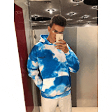 a man in a blue tie dye hoodie is taking a selfie in a bathroom mirror .