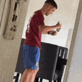a man in a maroon shirt and blue shorts is standing in a living room holding a can of soda .