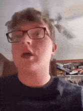 a young man wearing glasses and a black shirt is standing in a room with a ceiling fan .