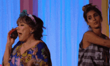 two women are standing next to each other on a stage . one is talking on a cell phone while the other is dancing .