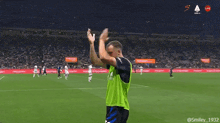 a man in a green vest applauds on a soccer field in front of a sign that says live