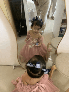 a little girl in a pink dress looking at herself in a mirror