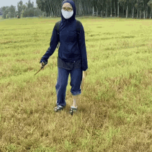 a woman wearing a mask and a blue jacket is standing in a field holding a pair of scissors .