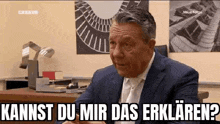a man in a suit and tie is sitting at a desk and talking to someone .