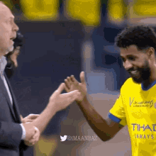 a man in a suit shakes hands with a soccer player in a yellow shirt that says tihal airways