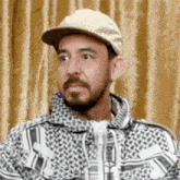 a man with a beard wearing a hat and a jacket is sitting in front of a curtain .