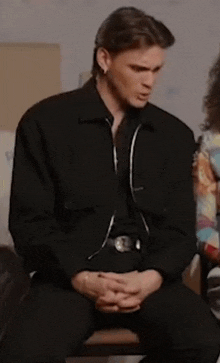 a man in a black jacket is sitting with his hands folded .