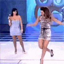 a woman in a silver dress is dancing on a stage while another woman sings into a microphone .