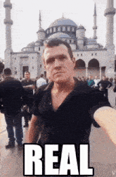 a man taking a selfie in front of a mosque with the word real on the bottom right