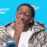 a man in a snakeskin jacket is sitting in front of a blue background and covering his mouth with his hand .