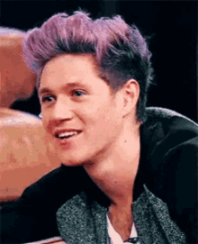 a young man with purple hair is smiling while wearing a black jacket .