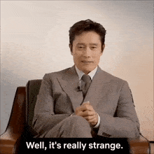 a man in a suit and tie is sitting in a chair with the words well it 's really strange behind him