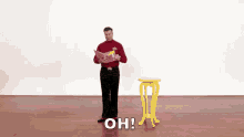 a man in a red shirt and black pants is standing next to a yellow table with the words oh written on it .