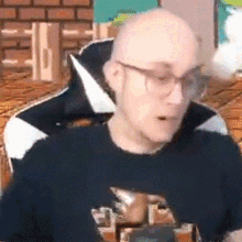 a bald man wearing glasses and a black shirt is sitting in a chair in front of a brick wall .
