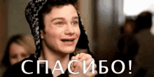 a young man wearing a hat is smiling and holding his hands up in front of a sign that says `` спасибо '' .