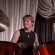 a man wearing a black tank top and a cross necklace smiles in front of a drum set