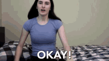 a woman in a blue shirt is sitting on a bed saying okay .