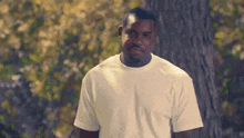 a man in a white t-shirt is standing in front of a tree and looking at the camera .