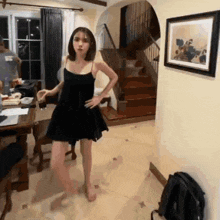 a woman in a black dress is standing in a living room next to stairs