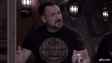 a man with a beard is sitting at a table holding a water bottle and asking who are you .