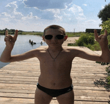 a shirtless boy wearing sunglasses stands on a wooden pier