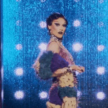 a drag queen is standing on a stage in front of a blue curtain with lights behind her .