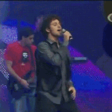 a man singing into a microphone while another man plays a guitar in the background