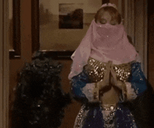 a woman wearing a pink veil and a belly dancer costume is standing in a hallway with her hands folded .