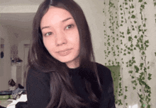 a woman with long dark hair looks at the camera in front of a wall with vines hanging from it