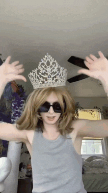 a woman wearing a crown and sunglasses holds her hands up in the air