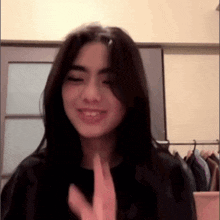 a woman with long black hair is smiling and waving at the camera while wearing a black shirt .