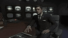 a man in a suit and tie sits at a desk in front of a wall of televisions with the letters cb on them