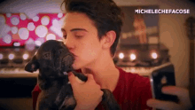 a young man is kissing a black dog on the cheek while looking at his phone .