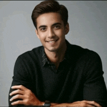 a man wearing a black sweater and a watch smiles with his arms crossed
