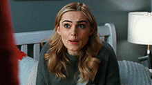 a woman in a grey sweater is sitting on a bed looking surprised .