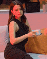 a woman in a black dress sits on a couch holding a cup