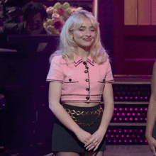a blonde woman in a pink top and black skirt smiles