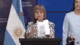 a woman stands at a podium holding a roll of paper