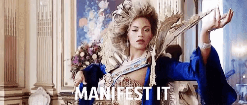a woman in a blue dress and wig is standing in a room with the words manifest it written on the screen .