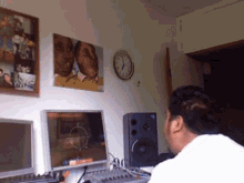 a man sits at a desk in front of two monitors and a clock with a picture of a couple on the wall behind him
