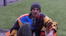 a man wearing a beanie and a colorful jacket is sitting in a chair on the grass .