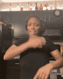 a woman in a black shirt is dancing in a kitchen with bottles on the shelf behind her
