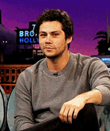 a man in a grey sweater is sitting in front of a sign that says bro holly