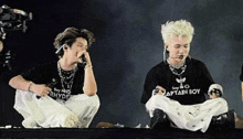 a man wearing a shirt that says captain boy sits next to another man on a stage .