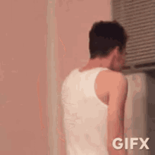 a man in a white tank top is standing in front of a window and looking down .