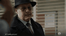 a man wearing a hat and sunglasses stands in front of a window with the nbc logo on the bottom right
