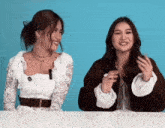 two women are sitting at a table with a blue background and laughing .