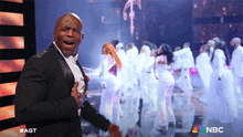 a man in a tuxedo is singing in front of a group of dancers and a nbc logo